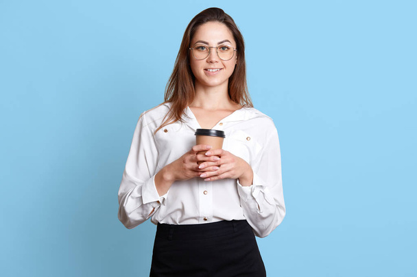 Studio shot of relaxed and smiling european woman with dark long hair and glasses, wearing blouse and skirt, holding takeaway coffee, enjoying great working day isolated over blue background. - Zdjęcie, obraz