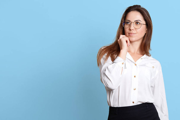 Portrait of young pensive woman touching her chin, has something in mind, posing with thoughtful facial expressions, wearing white shirt and black skirt, isolated over blue background. Copy space. - Foto, afbeelding