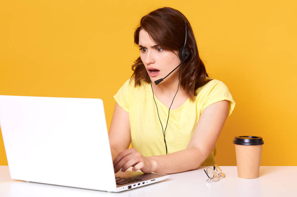 Half lengh photo of nervous young woman operator sitting with opened mouth, looking at laptopon screen, isolated over yellow background in studio. Dark haired surprised model working in office. - 写真・画像