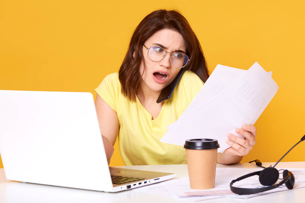 Photo of tense young brunette business woman working with paper documents in office isolated over yellow background, checking reports and finding lots of mistakes, has astonished expression. - Foto, immagini