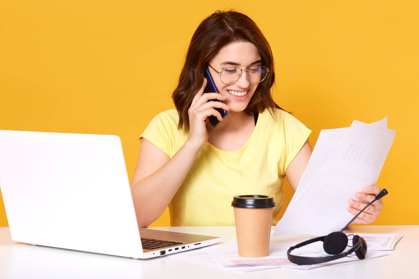 Portrait of woman talking on smartphone and holding papers. Lady calls on mobile phone, works in office, talks with client isolated over yellow background. Communication and consulting concept. - Photo, image