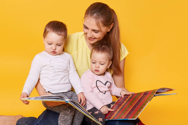 Studio shot of happy family: mother and little twins girls sitting on floor, reading books and viewing bright interesting pictures, spending time with mommy, selebrating Mother's Day. Children concept - Foto, Imagen