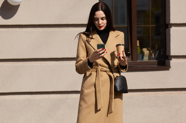 Image of serious young woman wearing elegant beige coat using her phone in sunny city street and drinking takeaway coffee, cheking social network or email while going to office, reading message. - Zdjęcie, obraz