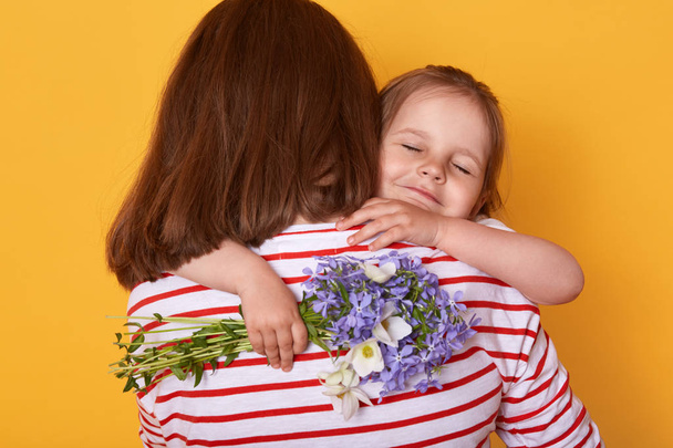 Happy Mother's day! Child daughter congratulates mom and gives her flowers. Mum and little girl hugging, charming kid closes eyes while enjoying moments. Family, holiday and togetherness concept. - Foto, Bild