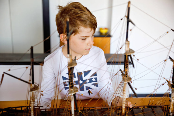 Little blond school kid boy playing with sailing ship model indoors. Excited child with yacht having fun after school at home. Happy boy building and creating toys - Photo, Image