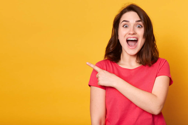 Indoor shot of positive attractive female with dark hair, dressed casually, points aside with index finger, has cheerful expression, shows something amazing at blank space, isolated on yellow wall. - Foto, afbeelding