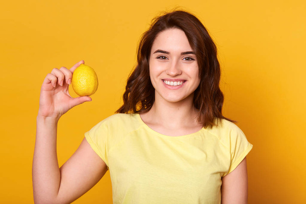Close up portrait of young beautiful dark haired woman with toothy smile, posing with lemon in hand, looking directly at camera with happy expression. Healthy food, vitamins and minerals concept. - Fotoğraf, Görsel