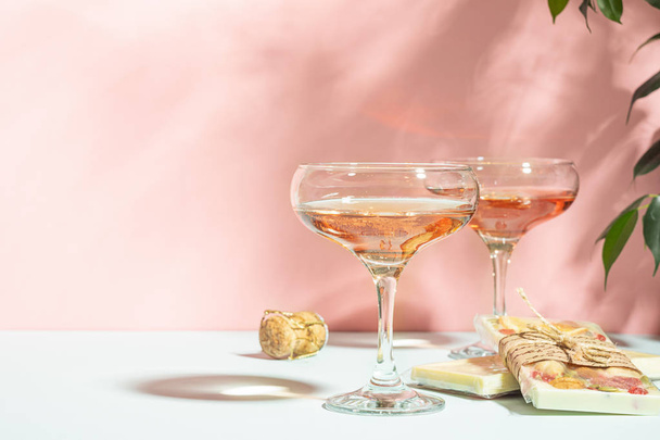 Champagner oder Wein in zwei eleganten Gläsern und einer Tafel weißer Schokolade trinken. sanft rosa Hintergrund helles Sonnenlicht. - Foto, Bild