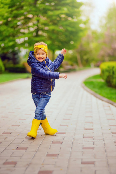 Niña jugando después de la lluvia en el parque. Una linda niña vestida con botas de goma amarillas chaqueta azul oscuro y jeans en la cabeza un pañuelo amarillo está atado. Caminar por la pasarela
 - Foto, Imagen
