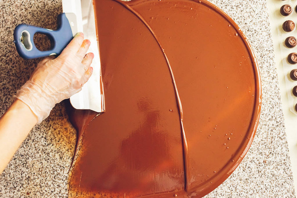 Φτιάχνω χειροποίητες σοκολάτες. Ο ζαχαροπλαστητής ρίχνει υγρή μαύρη σοκολάτα στην επιφάνεια. Γκρο πλαν. Επιλεκτική εστίαση. - Φωτογραφία, εικόνα