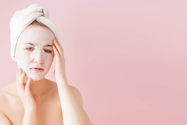 Beautiful young woman is applying a cosmetic tissue mask on a face on a pink background. Healthcare and beauty treatment and technology concept - Фото, изображение
