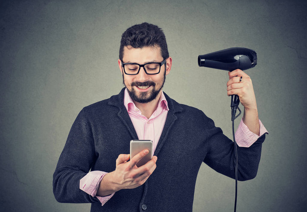Happy man reading news on smartphone blowing hair with hairdryer - 写真・画像