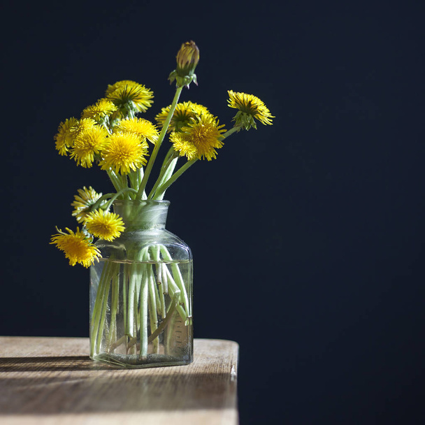 bouquet of yellow dandelions in a glass transparent jar on a wooden table against the background of a dark blue wall. - Фото, изображение