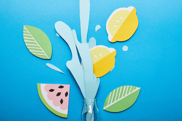 top view of bottle with paper cut water splash, lemons, leaves and watermelon on blue background - Photo, Image