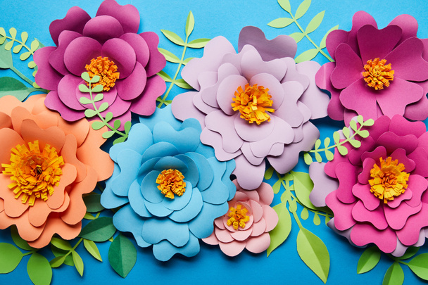 top view of bright colorful paper cut flowers with green leaves on blue background - Photo, Image