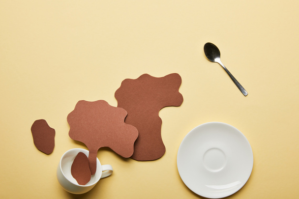 top view of paper cut coffee spills near white cup, saucer and spoon on beige background - Photo, Image