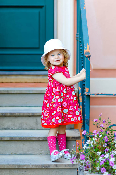 Portrait of beautiful little toddler girl in pink summer look clothes, fashion dress, knee socks and hat. Happy healthy baby child posing infront of colorful house. - Photo, Image