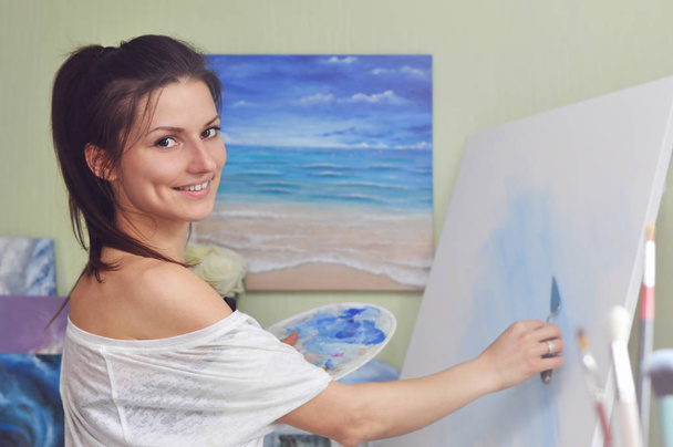 Una hermosa artista mujer pinta un cuadro de pinturas acrílicas azules sobre un gran lienzo blanco en su casa sobre el fondo de su pintura del mar
. - Foto, imagen