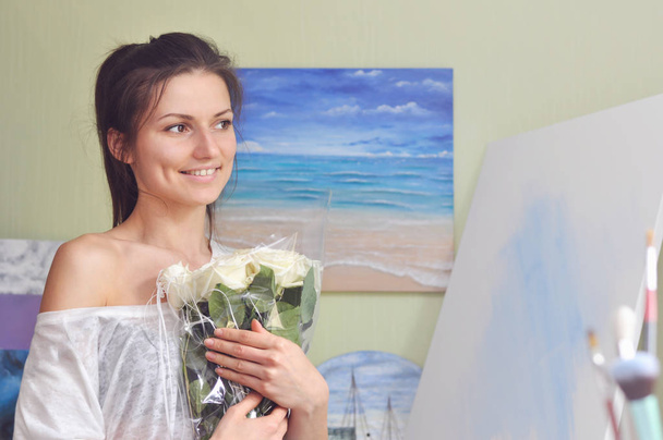 Ένας όμορφος ζωγράφος κρατά ένα μπουκέτο από λευκά τριαντάφυλλα στο σπίτι με φόντο μια εικόνα της θάλασσας. - Φωτογραφία, εικόνα