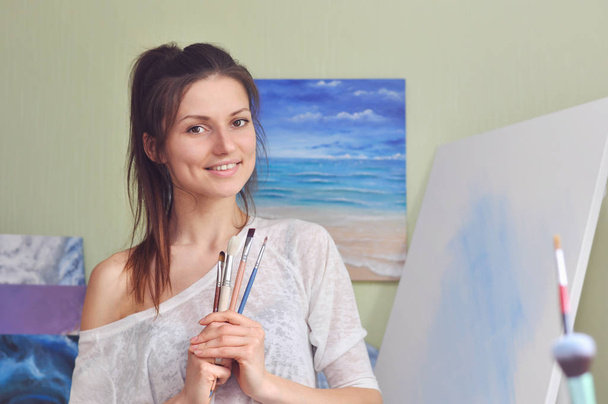 Piękny malarz posiada wiele różnych pędzli do malowania stojąc w pobliżu płótna w domu na tle malarstwa morskiego. - Zdjęcie, obraz