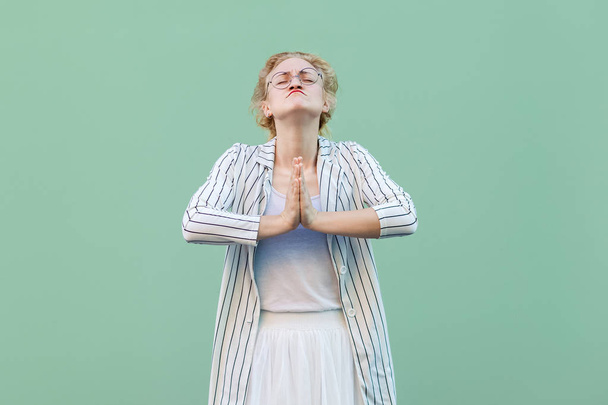 hoffnungsvolle junge blonde Frau in weiß gestreifter Bluse mit Brille steht mit Handflächen und betet auf hellgrünem Hintergrund. - Foto, Bild