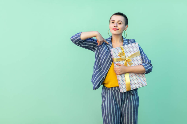 confident short hair beautiful hipster woman in yellow shirt and striped suit pointing finger to herself and holding present box with proud face on green background - Fotó, kép
