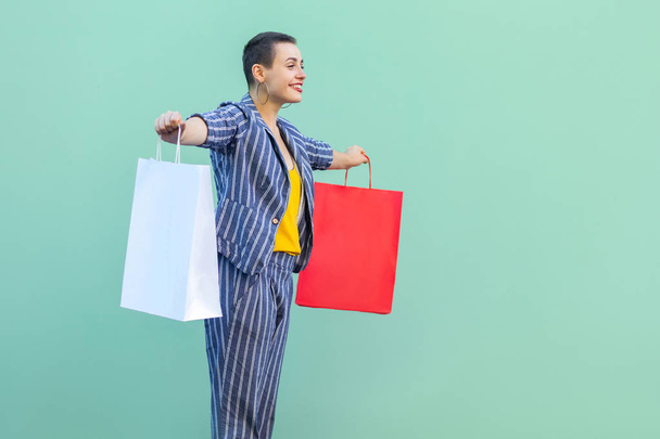 Side view of satisfied short hair beautiful hipster woman in yellow shirt and striped suit holding shopping bags with raised arms and smiling on green background - Foto, imagen