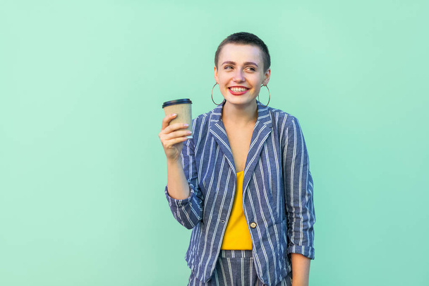positive resting short hair beautiful hipster woman in yellow shirt and striped suit drinking coffee and smiling while looking away on green background  - Foto, imagen