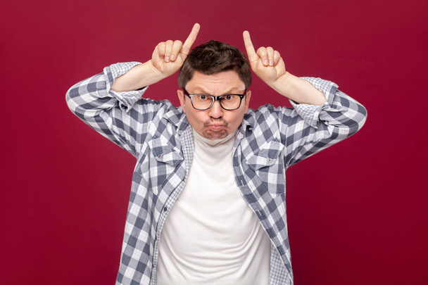 angry middle aged businessman in casual checkered shirt and eyeglasses standing with cow gesture horns on head and looking at camera on dark red background. - Valokuva, kuva