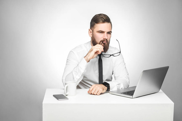 Thoughful bearded young boss in white shirt and black tie sitting at table and taking off glasses while looking aside on grey background  - Фото, изображение