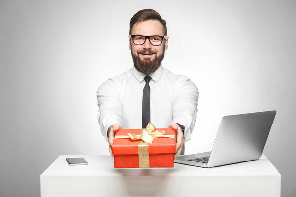 happy young boss in in white shirt and black tie sitting at table with laptop and holding red box on grey background - Zdjęcie, obraz