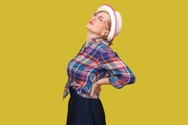 side view of modern stylish mature woman in casual style with hat standing and holding her painful back on yellow background. - Foto, afbeelding