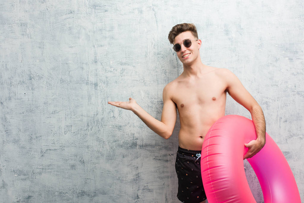 junger Mann hält einen rosafarbenen aufblasbaren Donut in einem Badeanzug und hält aufgeregt eine Kopie auf der Handfläche. - Foto, Bild