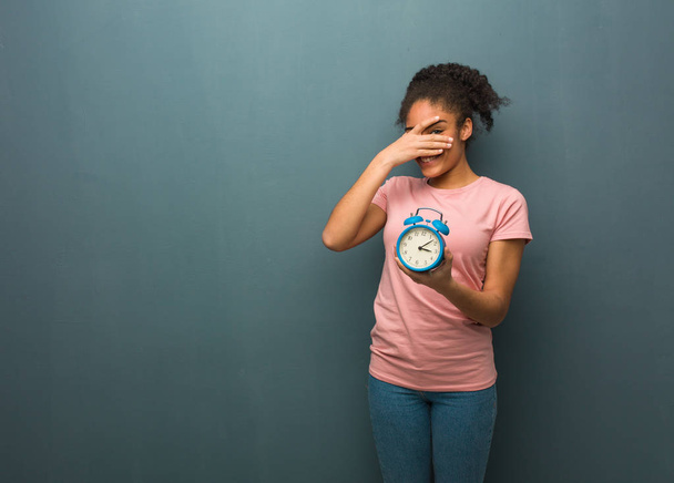 Young black woman embarrassed and laughing at the same time. She is holding an alarm clock. - Foto, Bild