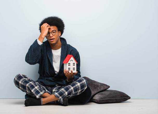 Joven hombre negro sosteniendo un modelo de casa sentado en el suelo preocupado y abrumado
 - Foto, imagen