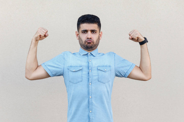 funny handsome young bearded man in blue shirt standing with raised arms and looking at camera with proud face on light beige wall background - Zdjęcie, obraz