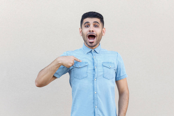 surprised handsome young bearded man in blue shirt pointing himself and looking at camera with amazed face on light beige wall background - Photo, Image