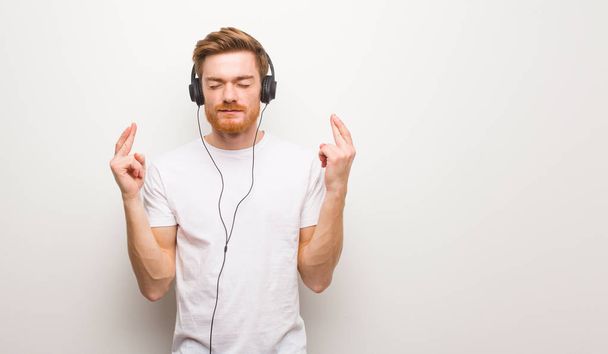 Jonge roodharige man kruist vingers voor geluk. Muziek beluisteren met hoofdtelefoon. - Foto, afbeelding