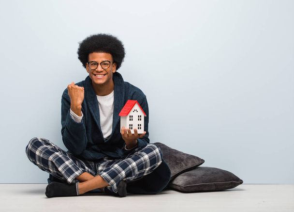 Joven hombre negro sosteniendo un modelo de casa sentado en el suelo sorprendido y sorprendido
 - Foto, imagen