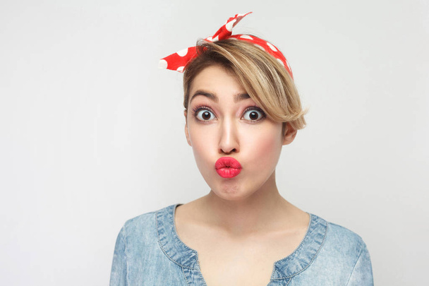 beautiful funny young woman in casual blue denim shirt with red headband looking at camera with big eyes and sending air kiss isolated on white background. - Photo, Image