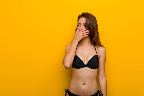 Joven mujer europea con bikini reflexivo mirando a un espacio de copia que cubre su boca con la mano
. - Foto, Imagen
