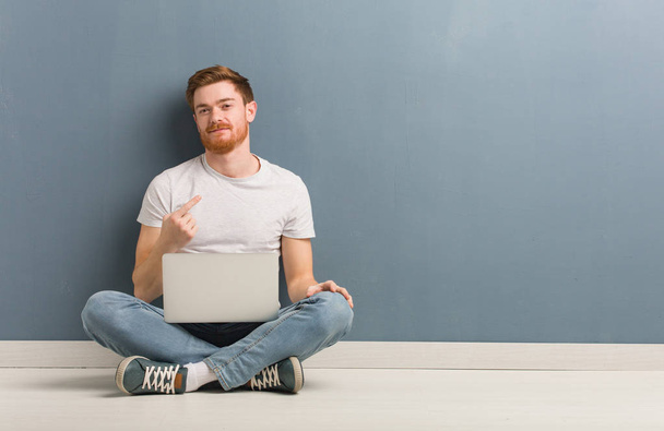 Young redhead student man sitting on the floor inviting to come. He is holding a laptop. - 写真・画像