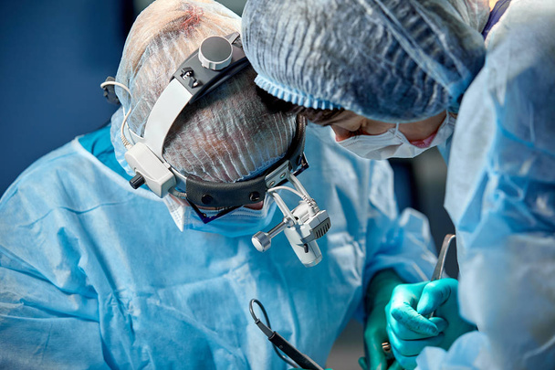 Cirurgião e seu assistente realizando cirurgia estética no centro cirúrgico do hospital. Cirurgião em máscara vestindo lupas durante a aquisição médica
. - Foto, Imagem