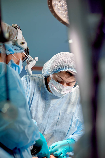 Хирург и его ассистент выполняют косметическую операцию в операционной больницы. Хирург в маске носит лупы во время медицинского осмотра
. - Фото, изображение