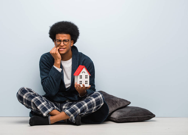 Joven hombre negro sosteniendo un modelo de casa sentado en el suelo mordiendo clavos, nervioso y muy ansioso
 - Foto, imagen