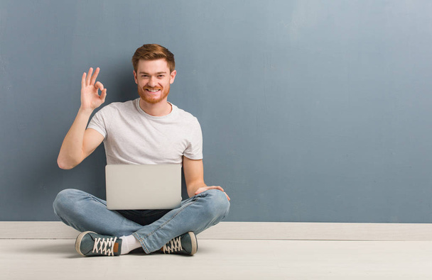 Jovem estudante ruiva homem sentado no chão alegre e confiante fazendo ok gesto. Ele está segurando um laptop
. - Foto, Imagem