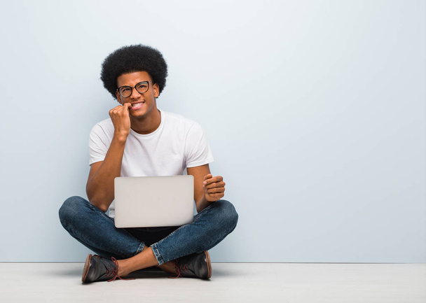 Giovane uomo nero seduto sul pavimento con un computer portatile che morde le unghie, nervoso e molto ansioso
 - Foto, immagini