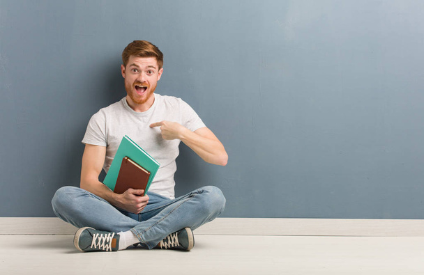 Young redhead student man sitting on the floor surprised, feels successful and prosperous. He is holding books. - 写真・画像