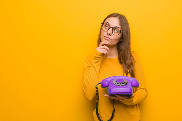 Young pretty caucasian woman doubting and confused. She is holding a vintage telephone. - Photo, Image