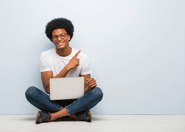 Jonge zwarte man zit op de vloer met een laptop lachend en wijzend naar de zijkant - Foto, afbeelding